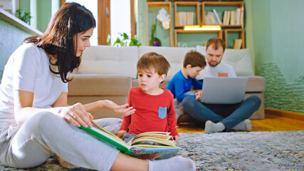 Fototapeta premium Big young family spending a day at home mom reading a story with her son dad with other kid doing some activities from the laptop