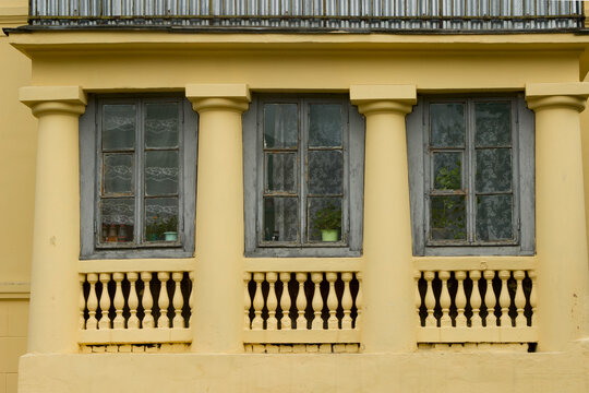 Three wooden window frames with cracked paint. The yellow facade of an old cozy European apartment building with columns, with geraniums and curtains in the windows.