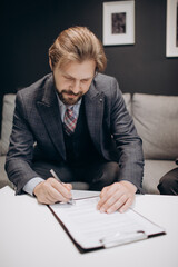 Mature bearded man in formal clothing signing documents while sitting on grey couch. Competent businessman starting financial relationship with new company.