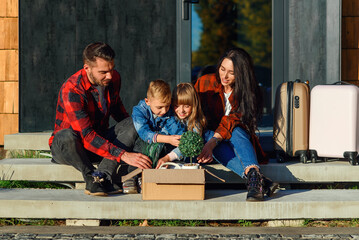 Obraz na płótnie Canvas Satisfied young parents with their happy children sitting on new house's stairs and get from the carton box green flower pots and clock. New stylish cozy home of lovely family near forest.