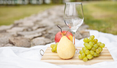 Picnic in the nature in the park. A glass of wine green grapes yellow pear red apple lilac on a wooden board on a white tablecloth in the grass in the garden on a stone path.