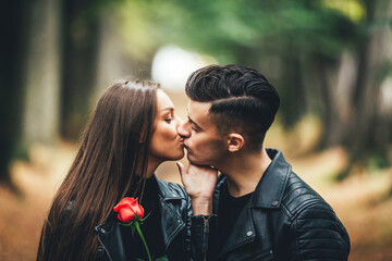 Happy girl with red rose kissing her boyfriend during their walk in park at the time of golden atunm sunset.