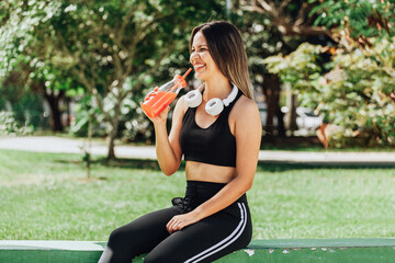 Woman wearing sportswear and headphones hydrating herself drinking juice after workout.