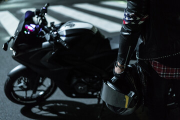 Piloto de moto con su moto detenido en la noche en la calle de la ciudad con una pañoleta en su brazo izquierdo sosteniendo su casco y su moto en el fondo de la toma