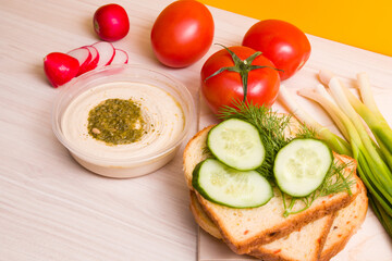 sliced cucumber on sliced bread, green onions, vegetables and a bowl with hummus on a wooden table, vegan sandwich for breakfast, proper nutrition concept