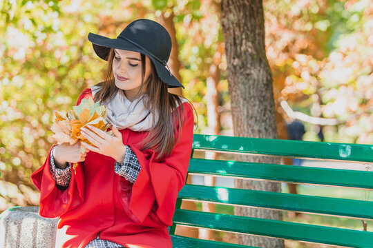 Closeup of romantic girl sitting on a bench in park, petting yellow autumn leaves and dreaming with pleased facial expression.