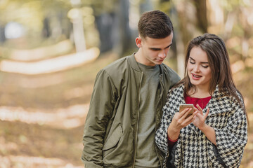 Obraz premium Copyspace photo of cheerful girl, showing photos to her boyfriend, while they are spending free time in autumn park.