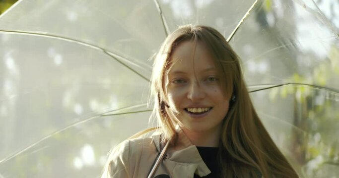 Anamorphic lens - Portrait of smiling young woman, looking into the camera and playing with transparent umbrella. Spring park. Anamorphic lens