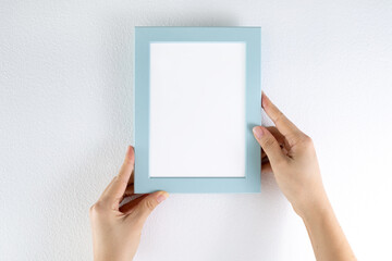 Female hand holding blank blue picture frame against a white wall.