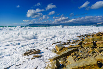 Storm in Tsemesskaya Bay. Turquoise waves with beautiful snow-white foam fall on Cape Doob. There are large yellowish chunks of rock on the shore. On the horizon Novorossiysk and the mountains