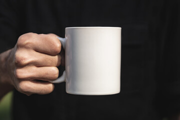 Man holding a white cup of hot beverage outdoors. Man's hand holding a blank mug against black background. Close up, mockup image, copy space for text.