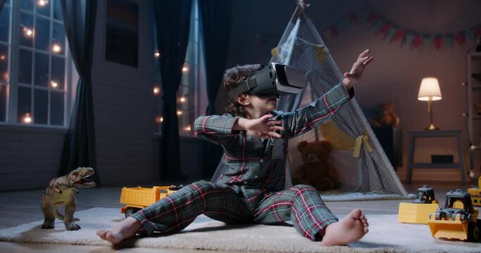 Cute little boy wearing pajamas is diving into virtual reality world before bedtime, using vr 3d headset, playing video games - modern technology concept 4k footage
