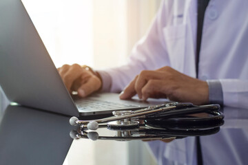 Male doctor in white coat working on laptop computer with medical stethoscope on the desk in...