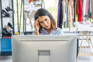 Young Asian woman business owner sitting at a desk in a clothing shop looking at a computer monitor with a stressed worry unhappy headache expression on her face.