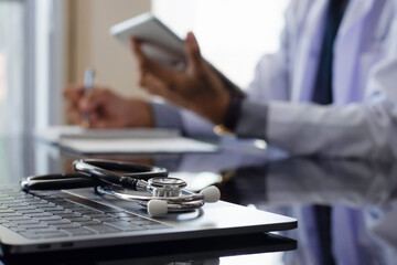 Male doctor in white coat, hand holding digital tablet and and writing note with laptop computer and medical stethoscope on the desk. Medical technology network concept.