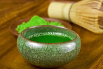 Green matcha tea in a cup with powdered matcha tea in a plate and bamboo matcha tea whisk also know as chasen on a wooden background.