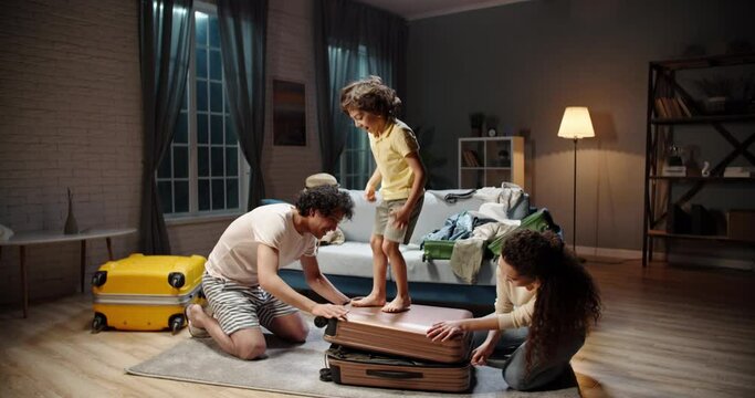 Young Asian Parents Are Trying To Close A Tightly Packed Suitcase While Their Funny Kiud With Curly Hair Is Dancing And Jumping On It. Family Preparing For Vacation 4k Footage