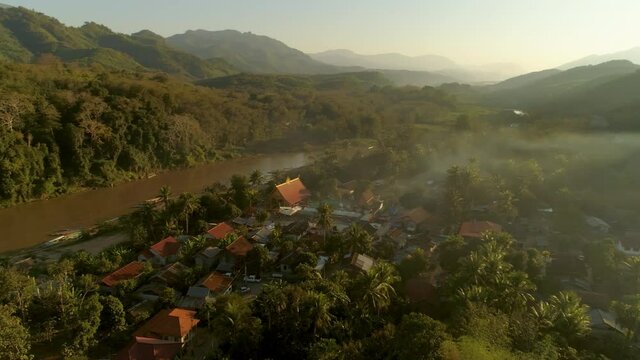 Aerial shot of houses in village amidst trees near river, drone panning over residential structures against sky during sunrise - Tat Sae Waterfalls, Laos