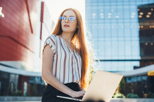 Busy caucasian entrepreneur with read hair and freckles wearing eyeglasses and working at the laptop outside - Powered by Adobe