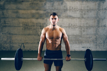 Fototapeta na wymiar Serious Caucasian shirtless man in shorts lifting barbell. In background wall.