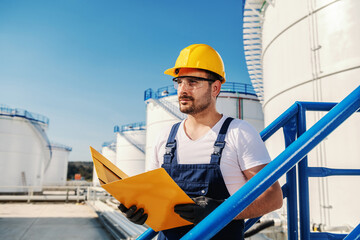 Young hardworking Caucasian unshaven refinery worker in working clothes with helmet on head and...
