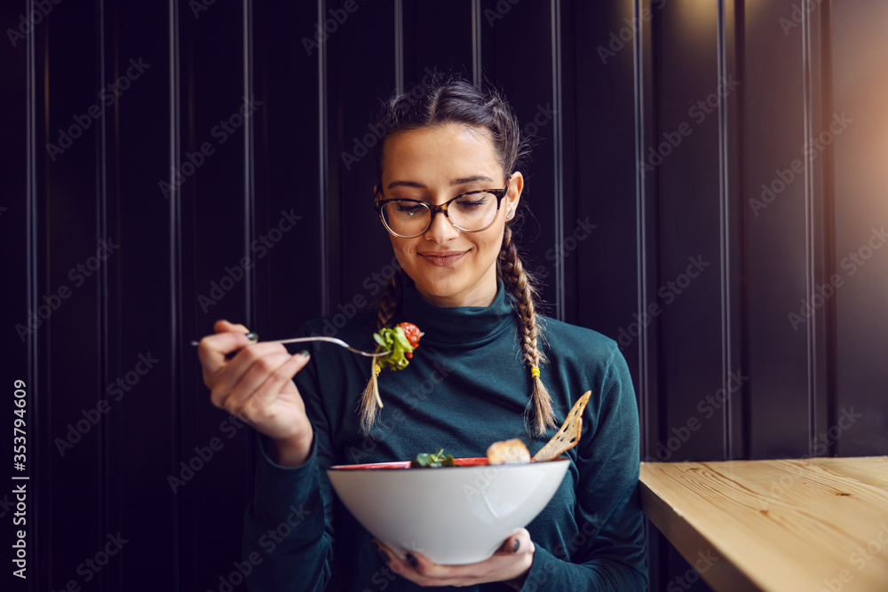 Wall mural young attractive teenage girl with healthy habits sitting in restaurant, holding bowl with salad and