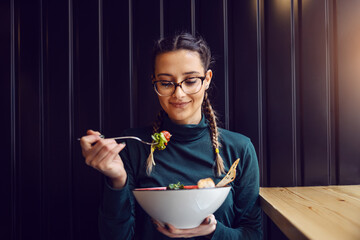 Young attractive teenage girl with healthy habits sitting in restaurant, holding bowl with salad...