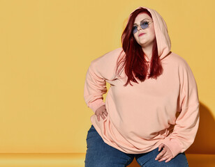 Young smiling red haired overweight woman in hoodie, jeans and sunglasses posing on floor with her...