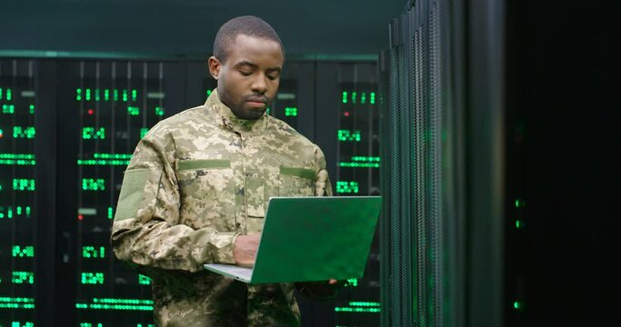 US army male African American officer in uniform stading at server wih secret data and using laptop computer to check the information. Military monitoring service room. Army networking concept.