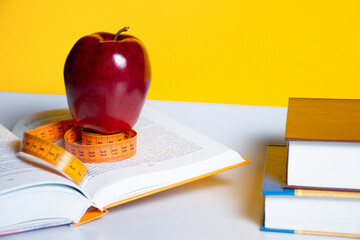 A ripe fresh red Apple stands on a measuring tape for measuring body volume and open scientific books about diet on a yellow background: concept of healthy eating against obesity, how to get in shape