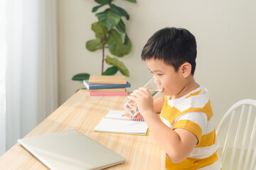 Naklejka premium Portrait of little Asian boy drinking water while using laptop, sitting at desk and doing homework. E-learning and education concept.