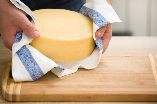 Seller’s hands are holding a cheese head wrapped in a towel closeup, over a chopping wooden board. Gourmet dairy products, food market