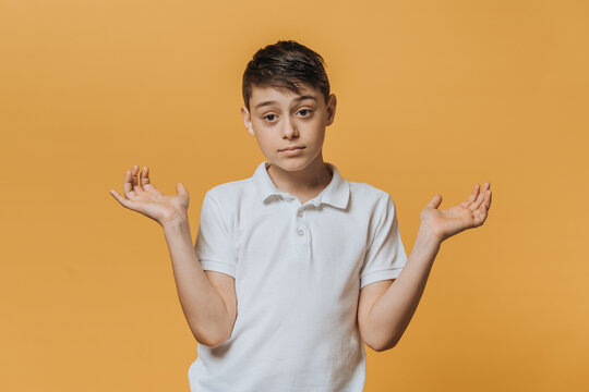 Disappointed Young Boy In A White T-shirt Spreads His Hands In Doubt Mood Over Yellow Studio Backdrop. Sincere People Emotions.