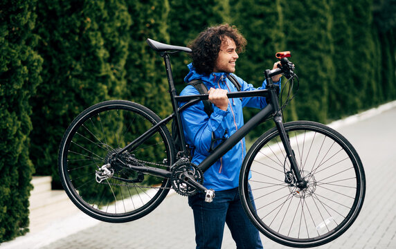 Outdoor image of handsome hipster man holding his bike on the shoulder next to the house. The male courier with curly hair delivers parcel cycling with a bicycle in the city. Delivery service concept.