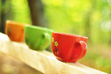 Three cups red, green and yellow stand on the railing of a wooden staircase in the forest.