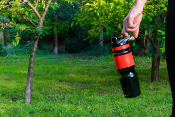 .Girl's hand holds a bottle on a background of green park. Black and red sports bottle of water.