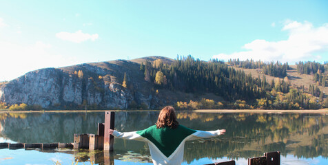 Young woman stands by the lake among mountains with arms outstretched. Panorama.