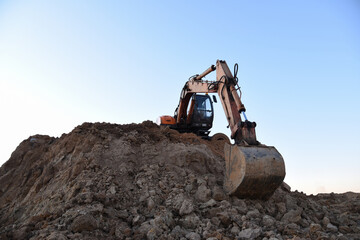 Obraz premium Bucked wheel excavator on earthmoving. Backhoe digs ground in sand quarry on blue sky background. Construction machinery for excavation, loading, lifting and hauling of cargo on job sites
