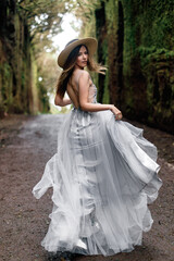 Fototapeta premium a young girl with long hair in a light dress and a straw hat runs into the forest and turns around