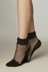 Cropped shot of female legs in black nylon sheer socks with black serrated pattern, black toes, bottoms and rims. The lady is standing on tiptoe on the gray background. 