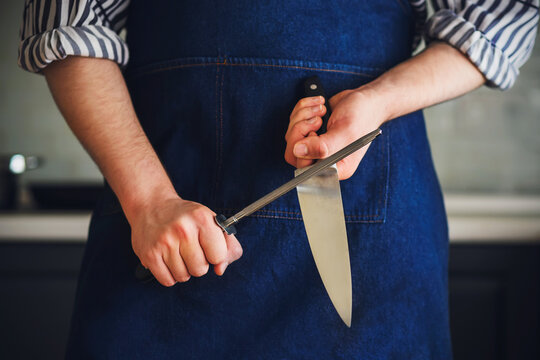 A man dressed in a blue kitchen apron and a striped shirt sharpens the blade of a knife with a whetstone before starting to cook. Home cooking. Kitchen utensils.