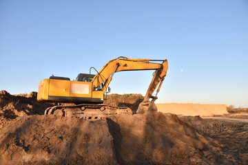Obraz premium Excavator working on earthmoving. Backhoe digs ground in sand quarry on blue sky background. Construction machinery for excavation, loading, lifting and hauling of cargo on job sites