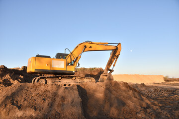 Excavator working on earthmoving. Backhoe digs ground in sand quarry on blue sky background. Construction machinery for excavation, loading, lifting and hauling of cargo on job sites