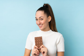 Young brunette woman over isolated blue background taking a chocolate tablet and happy