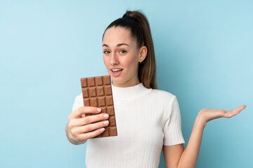Young brunette woman over isolated blue background taking a chocolate tablet and surprised