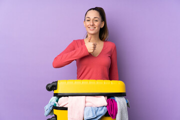 Traveler woman with a suitcase full of clothes over isolated purple background giving a thumbs up gesture