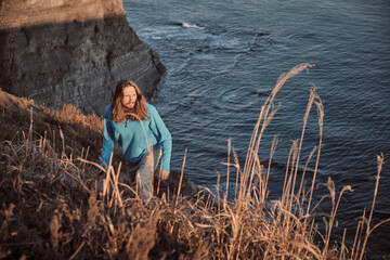 Young man with a long hair walking on an alpine meadow through the cliffs to the sea on a sunset with ocean background. Journey and outdoor concept.