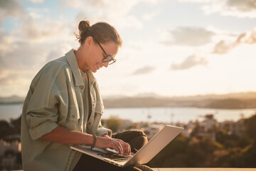 Beautiful young girl woman in eyeglasses sitting with a laptop on her balcony at sunset with a view...