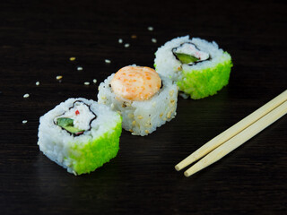 Three Japanese rolls at an angle, light chopsticks and sprinkled sesame seeds on a dark wood table.
