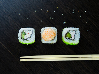Three Japanese rolls at an angle, light chopsticks and sprinkled sesame seeds on a dark wood table, top view.